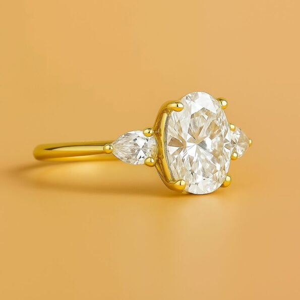 Oval cut three-stone diamond ring in yellow gold with pear side diamonds, side view on golden background.